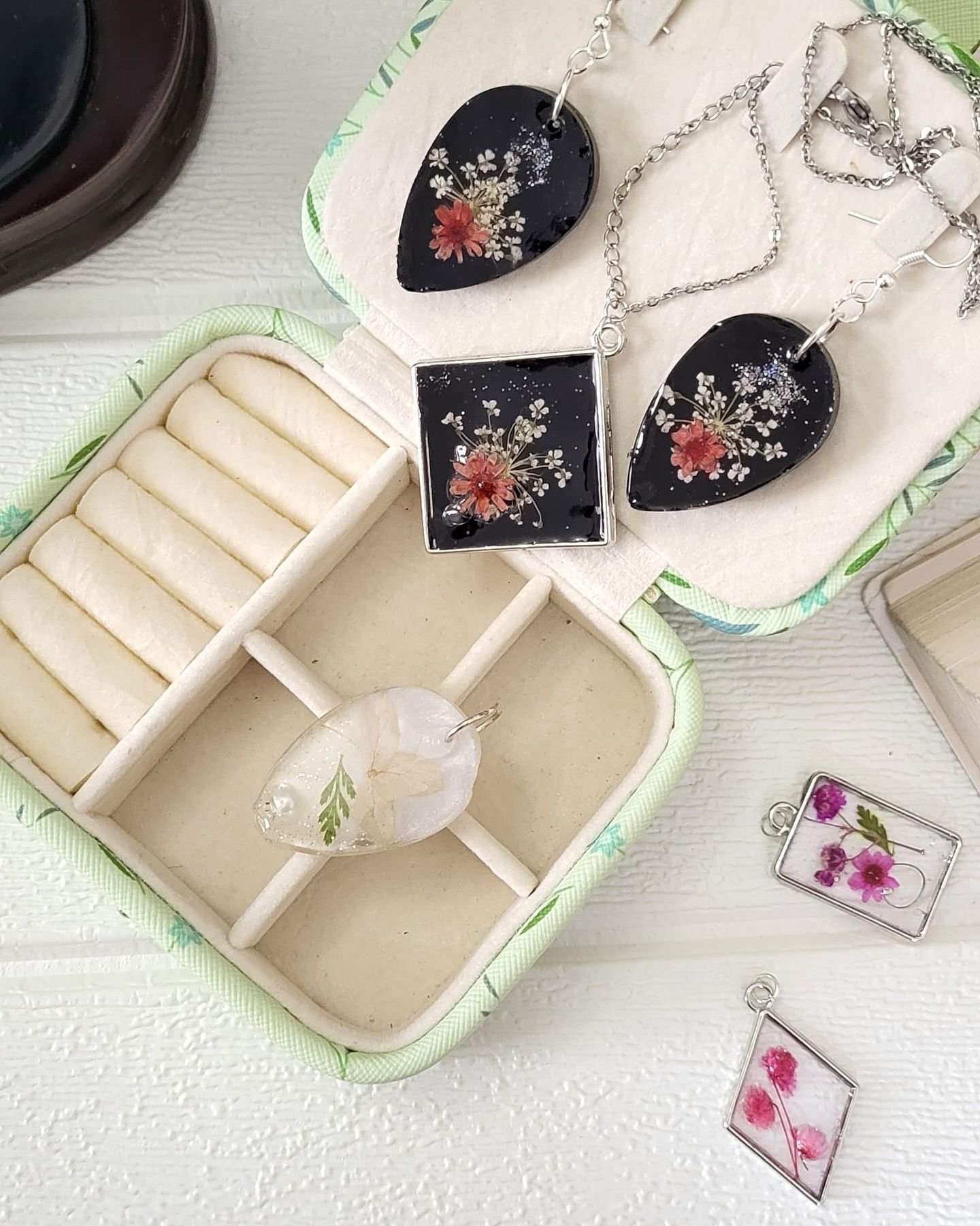 The set on the jewellery case seen from further back, a clear teardrop pendant of white petal and fern resting in the case beside it and two small pink-flowered pendants on the cloth at the right