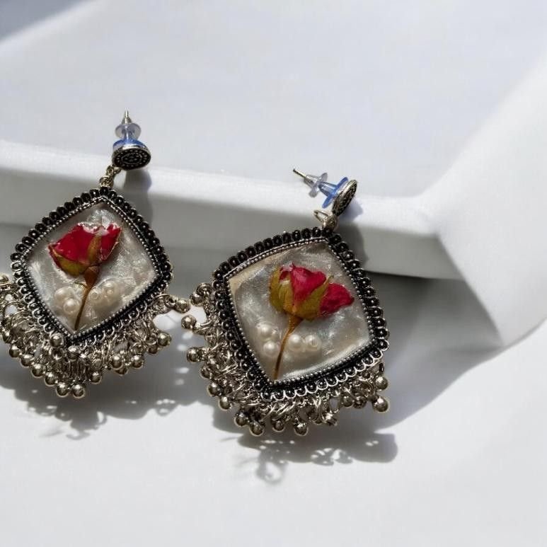 A pair of diamond-shaped earrings holding a red rose in pale resin, framed in ornate oxidised silver with a fringe of small silver bells, on a white surface
