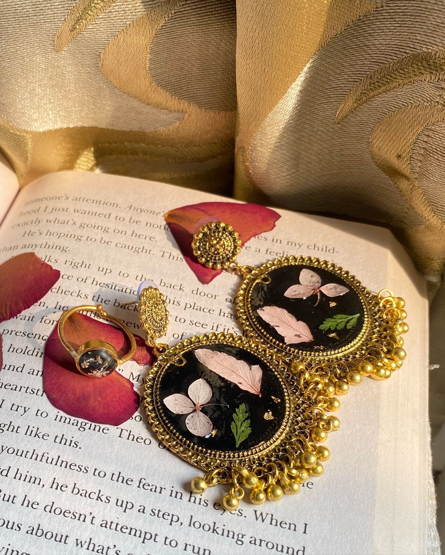 A pair of black resin jhumkas set with blush hydrangea petals and a green fern frond, framed in antique gold with a fringe of small gold bells, resting on the open pages of a book beside red rose petals