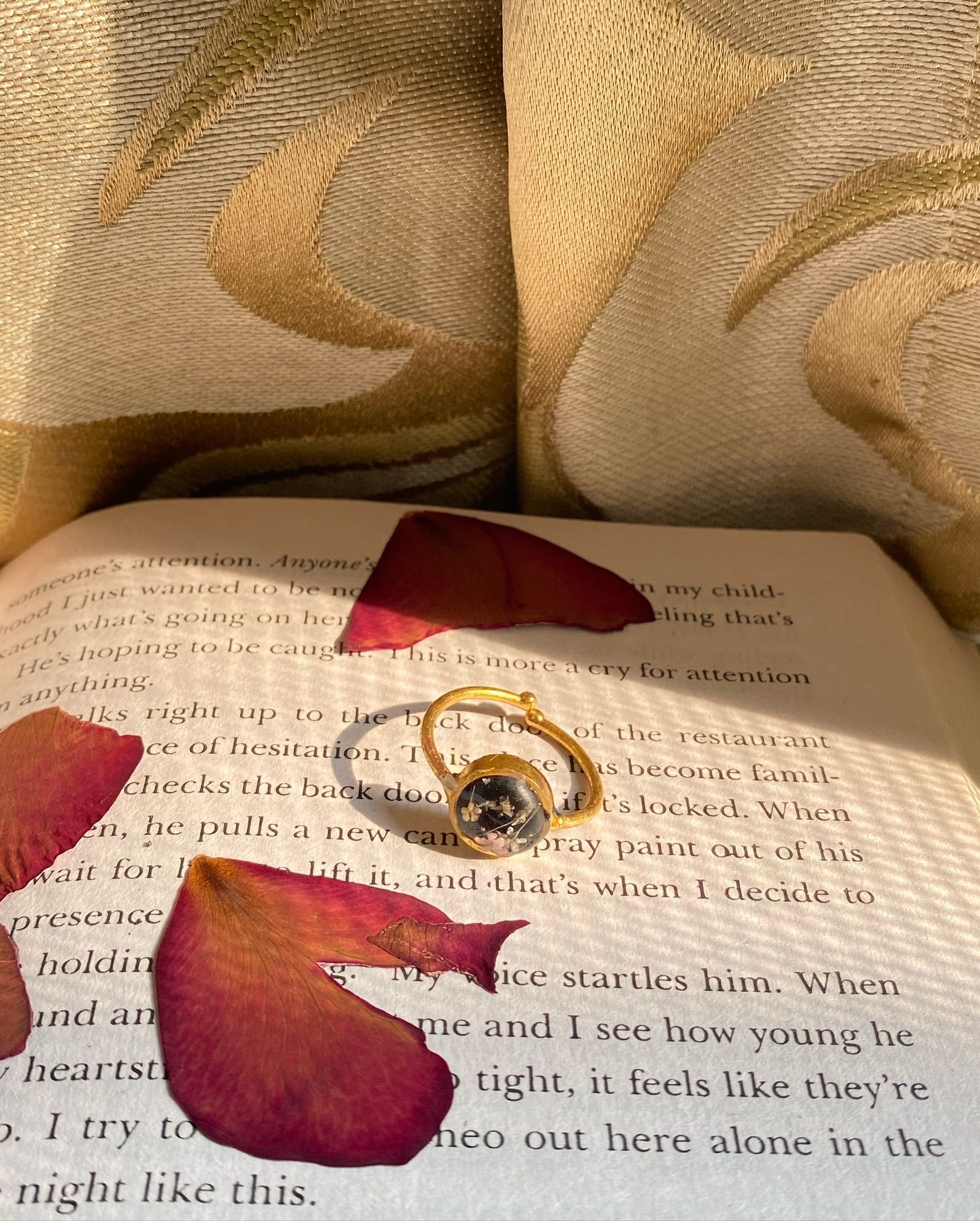 The matching ring — a slim gold band with a small dark resin stone — lying on the open book beside a red petal