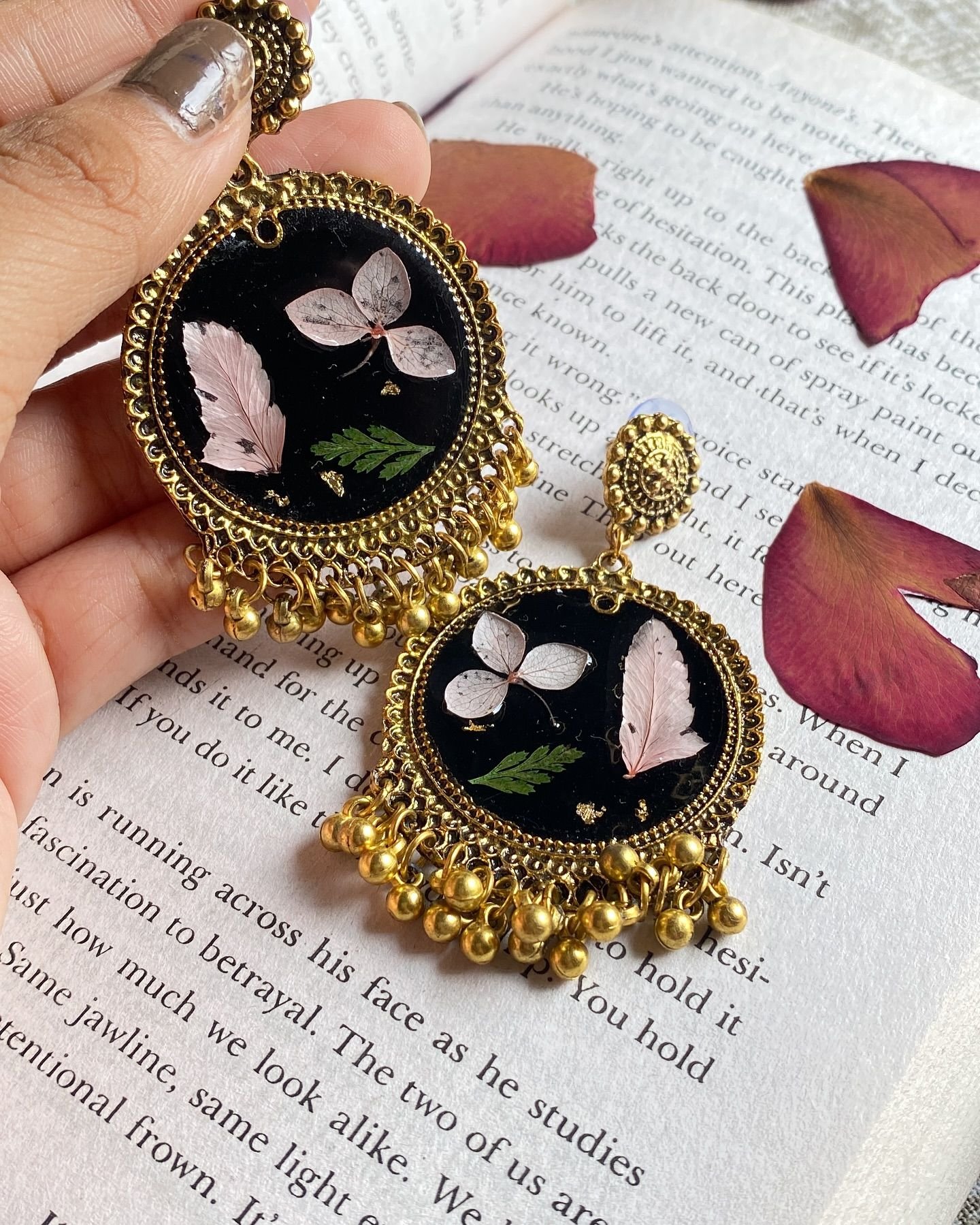 The jhumkas held between finger and thumb, close enough to show the pressed petals through the resin