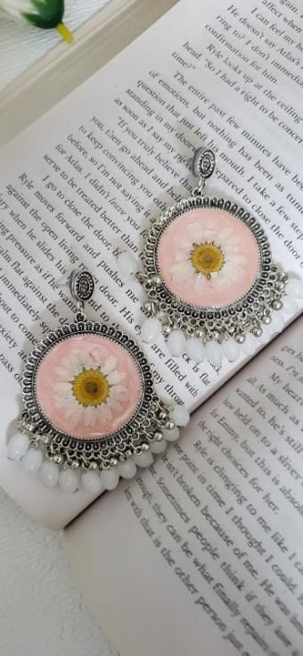 A pair of round oxidised silver jhumkas resting on the pages of an open book, each holding one white daisy with a yellow centre on a blush pink ground, with a fringe of white beads and small silver ghungroo, a green leaf at the top corner of the page