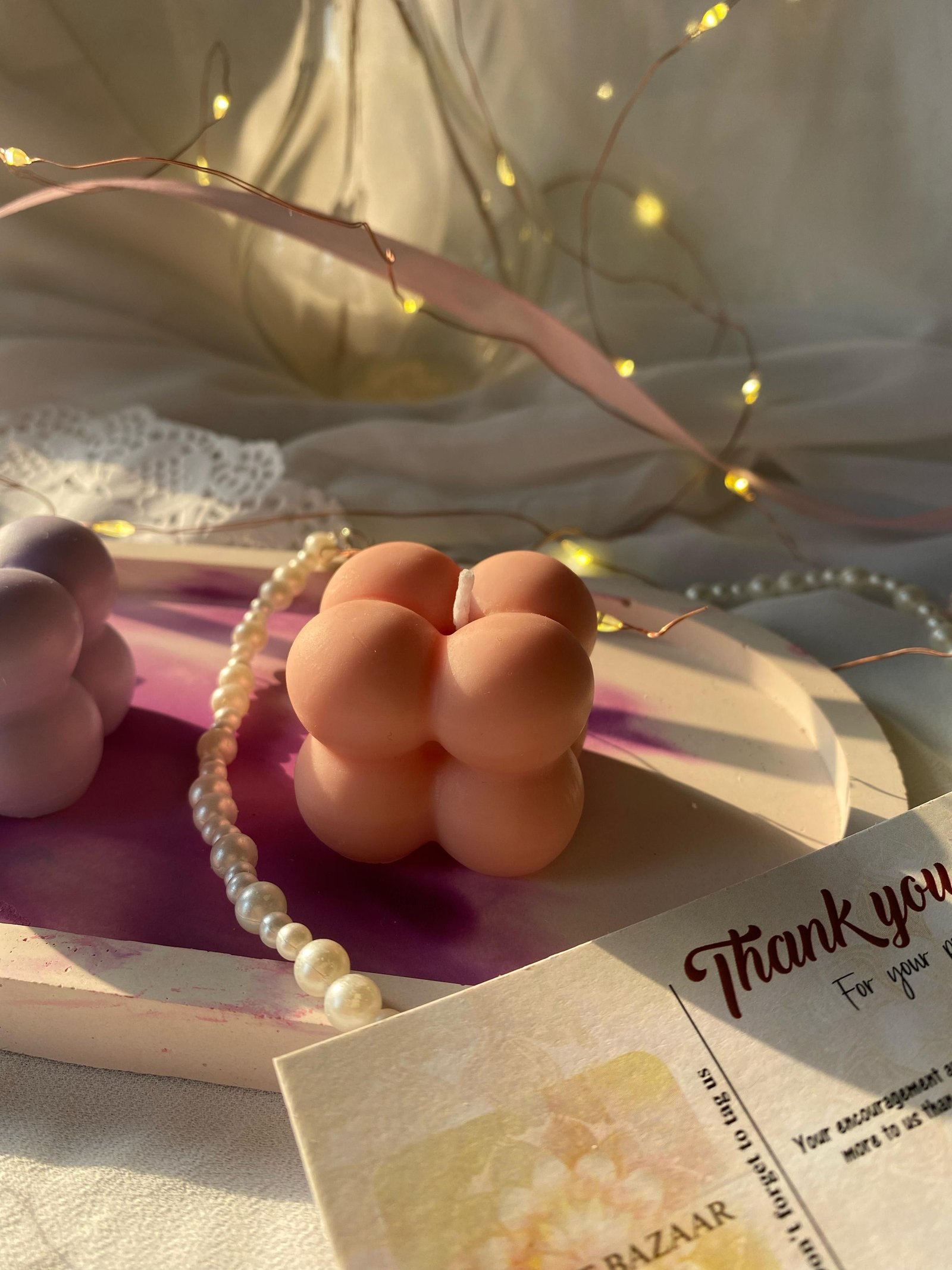 A peach-coloured bubble cube candle resting on a pink dish beside a string of pearls and a thank-you card