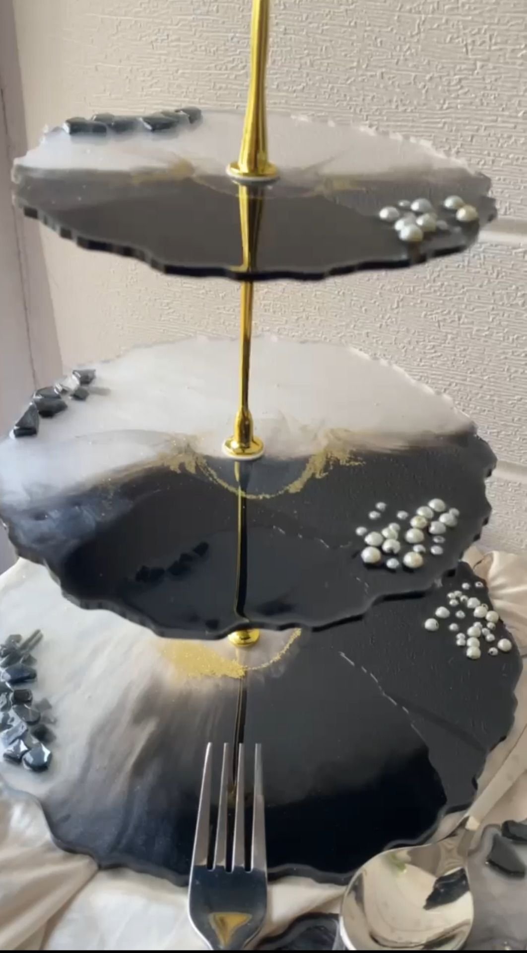 A three-tier cake stand, each tier a scalloped disc of black and white marbled resin veined with gold and set with clusters of pearls, threaded on a slim gold spindle, standing on a cloth-draped table with silver cutlery below