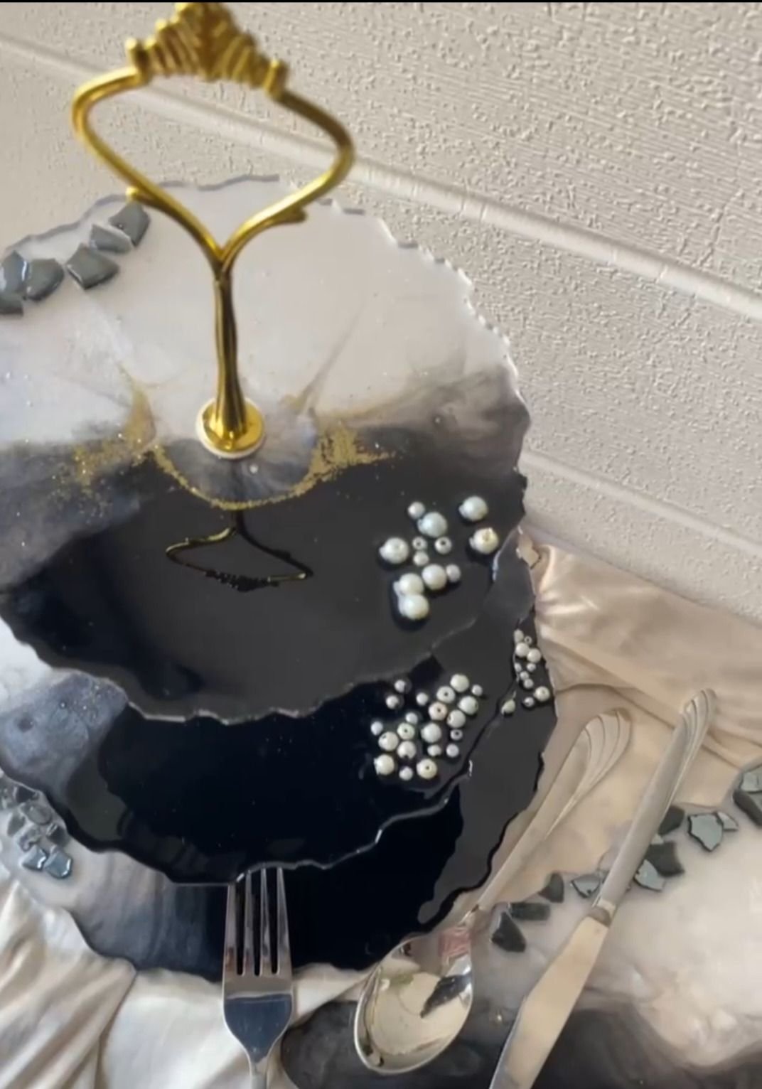 The cake stand seen closer from above, showing the ornate gold handle at its top, the gold veining running through the black and white resin and the pearls set into the surface