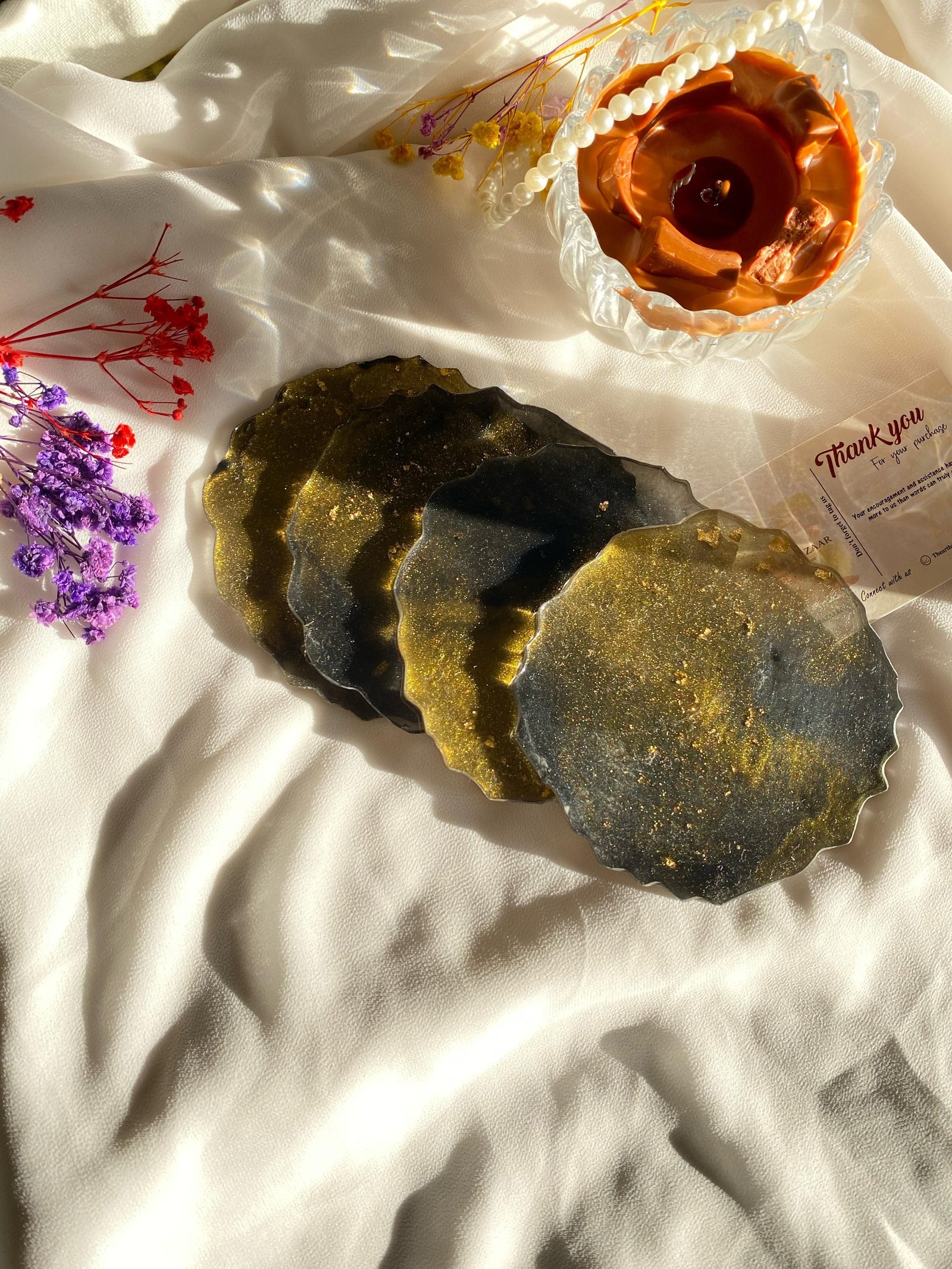 A stack of round coasters poured in dark green and black resin with gold veining, on white cloth beside dried flowers, with a copper dish and a thank-you card
