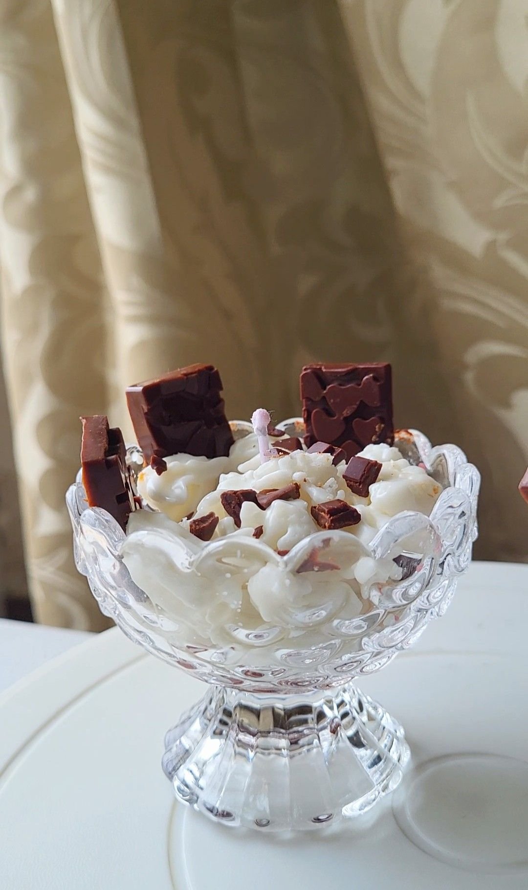 A candle made to look like a dessert: piped white wax cream topped with chocolate shards and a wafer, in a footed crystal glass