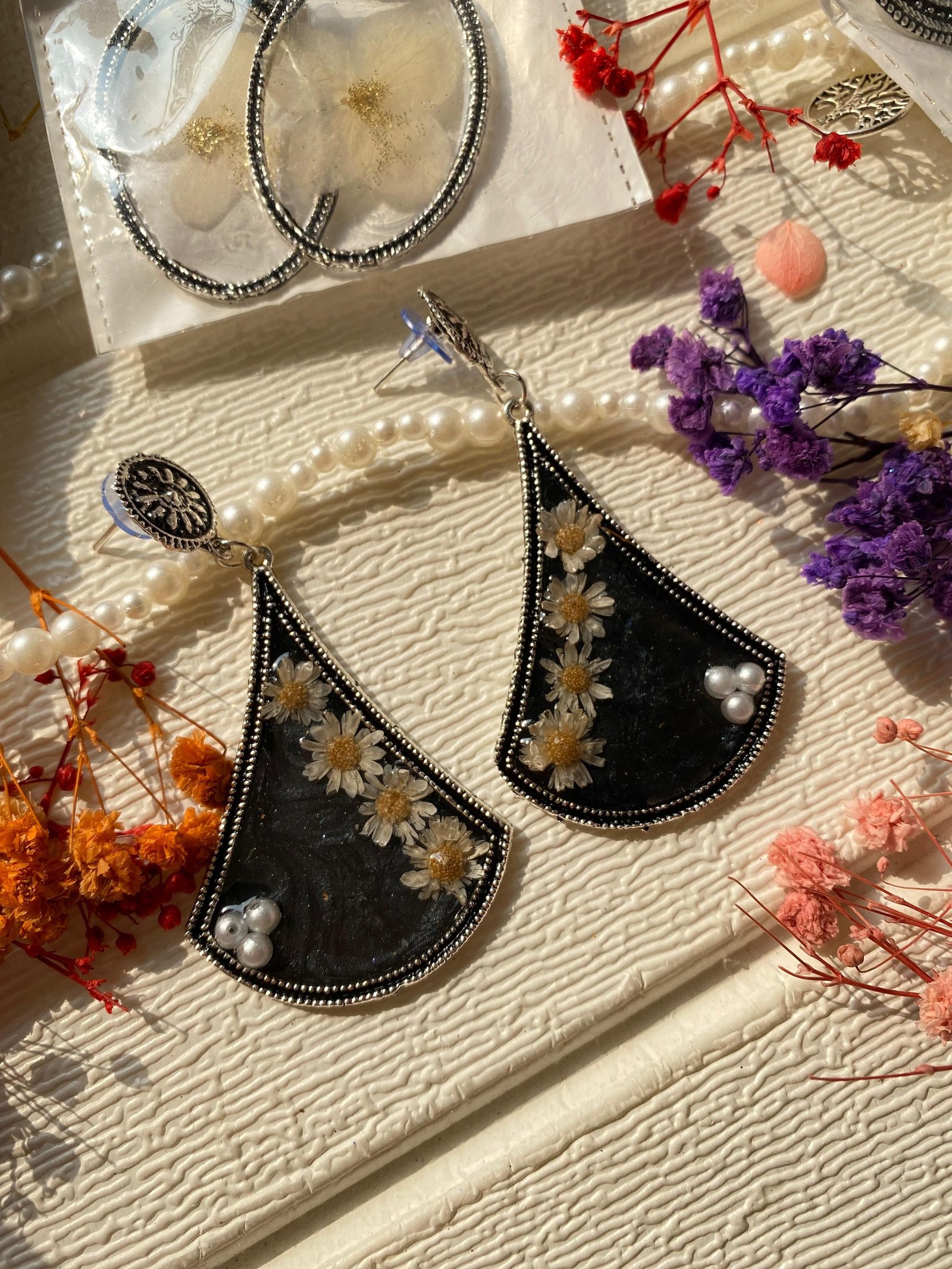 A pair of fan-shaped earrings in black resin scattered with small white flowers and a pearl, framed in oxidised silver, on textured cream cloth