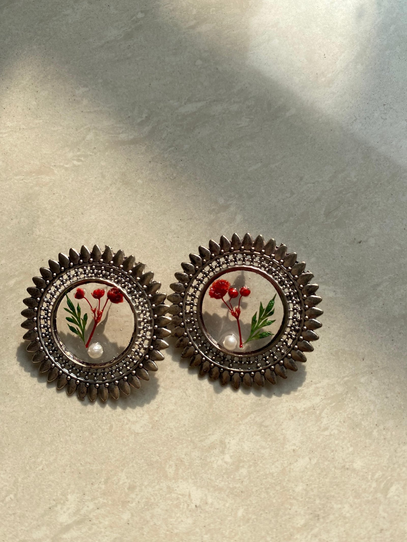 A pair of small round studs holding tiny red flowers and green leaves under clear resin, in ridged oxidised silver settings