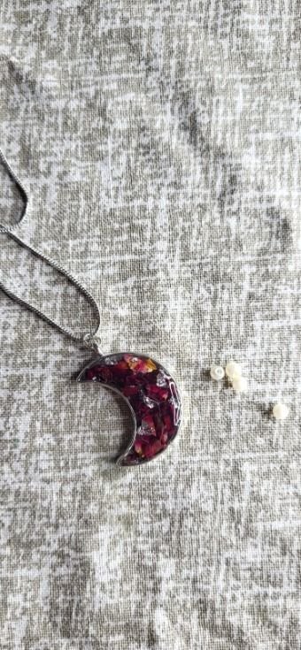 A crescent moon pendant in a fine silver frame, filled with deep magenta rose petals and flakes of gold, hanging from a slim chain laid out on grey-beige linen, two small pearl studs beside it