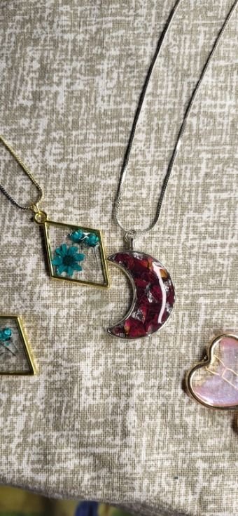 The same crescent on its chain beside a gold rhombus pendant holding one teal flower, a second gold rhombus below it and a pale pink shell-shaped pendant at the edge of the cloth