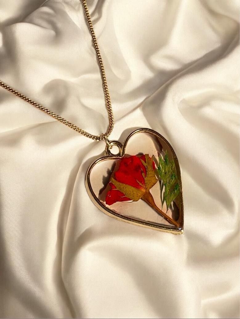 A heart-shaped pendant of clear resin holding a red rose and a green leaf, on a fine chain over cream satin