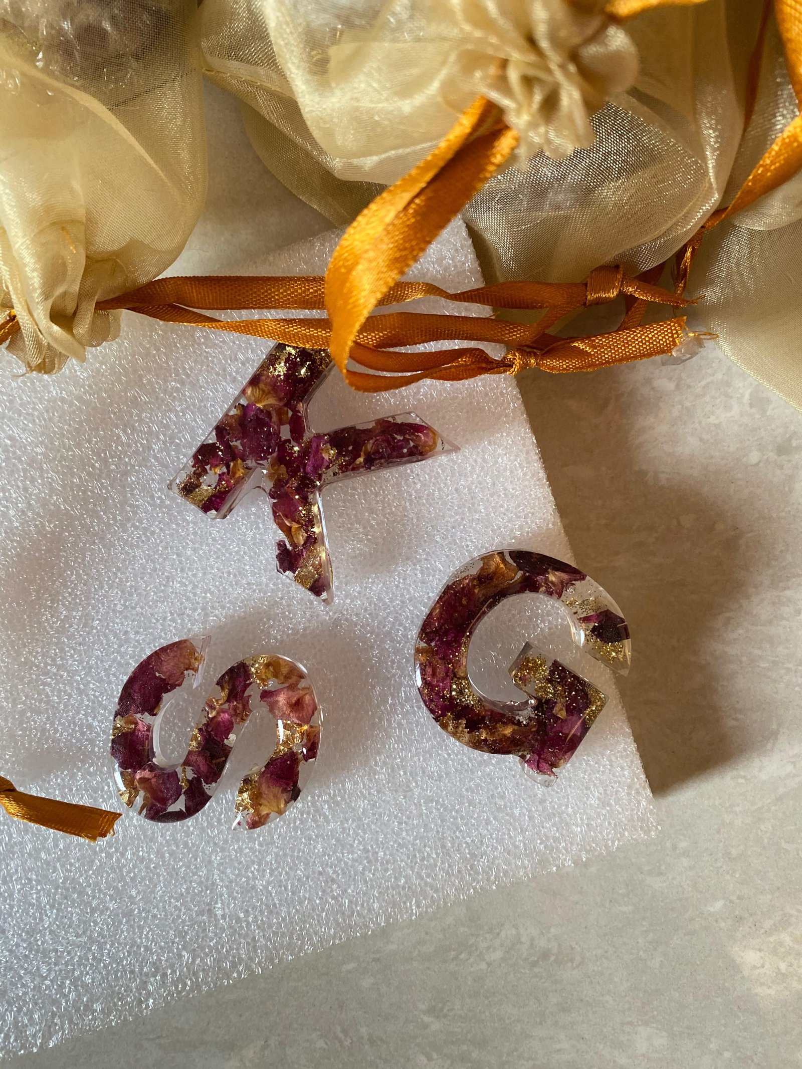 Three resin initials — K, S and G — each set with dark rose petals and flakes of gold leaf, laid on white foam with orange ribbons threaded through them and cream organza pouches behind