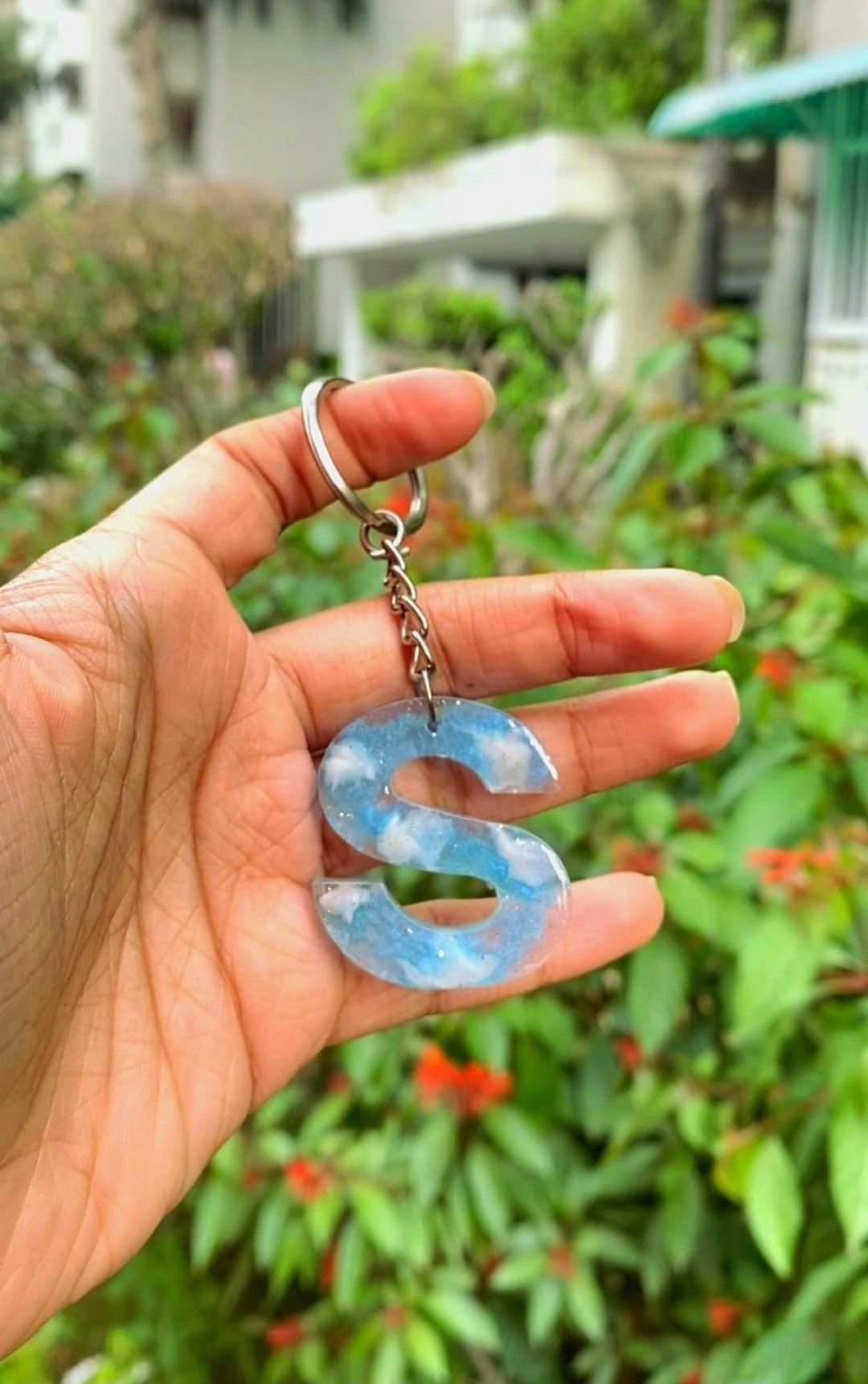 A resin keychain shaped as a single initial in pale blue, held up against a garden