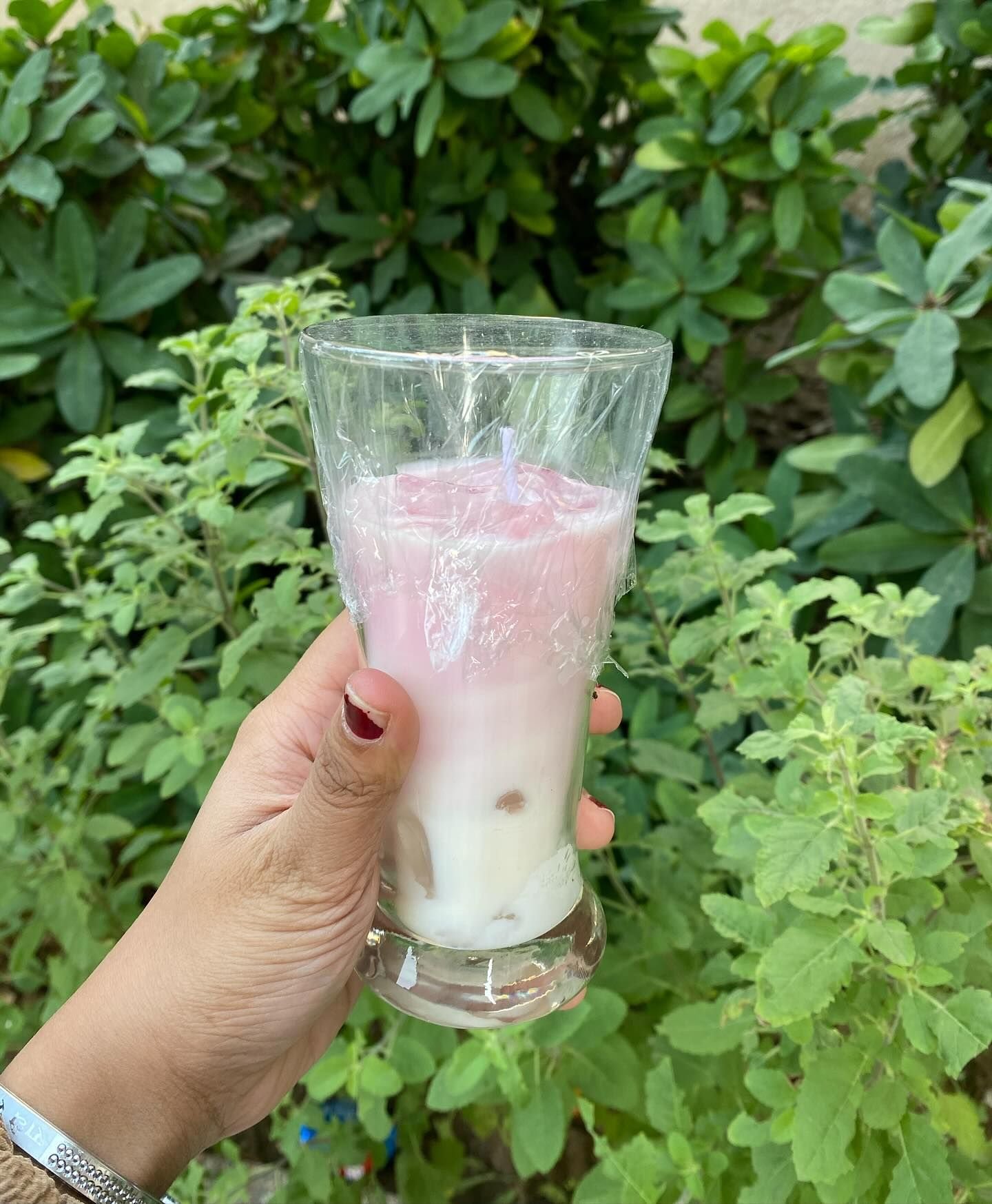 A candle poured to look like a latte in a tall glass — pink wax layered over cream, with the wick standing at the centre — held in the hand against green foliage