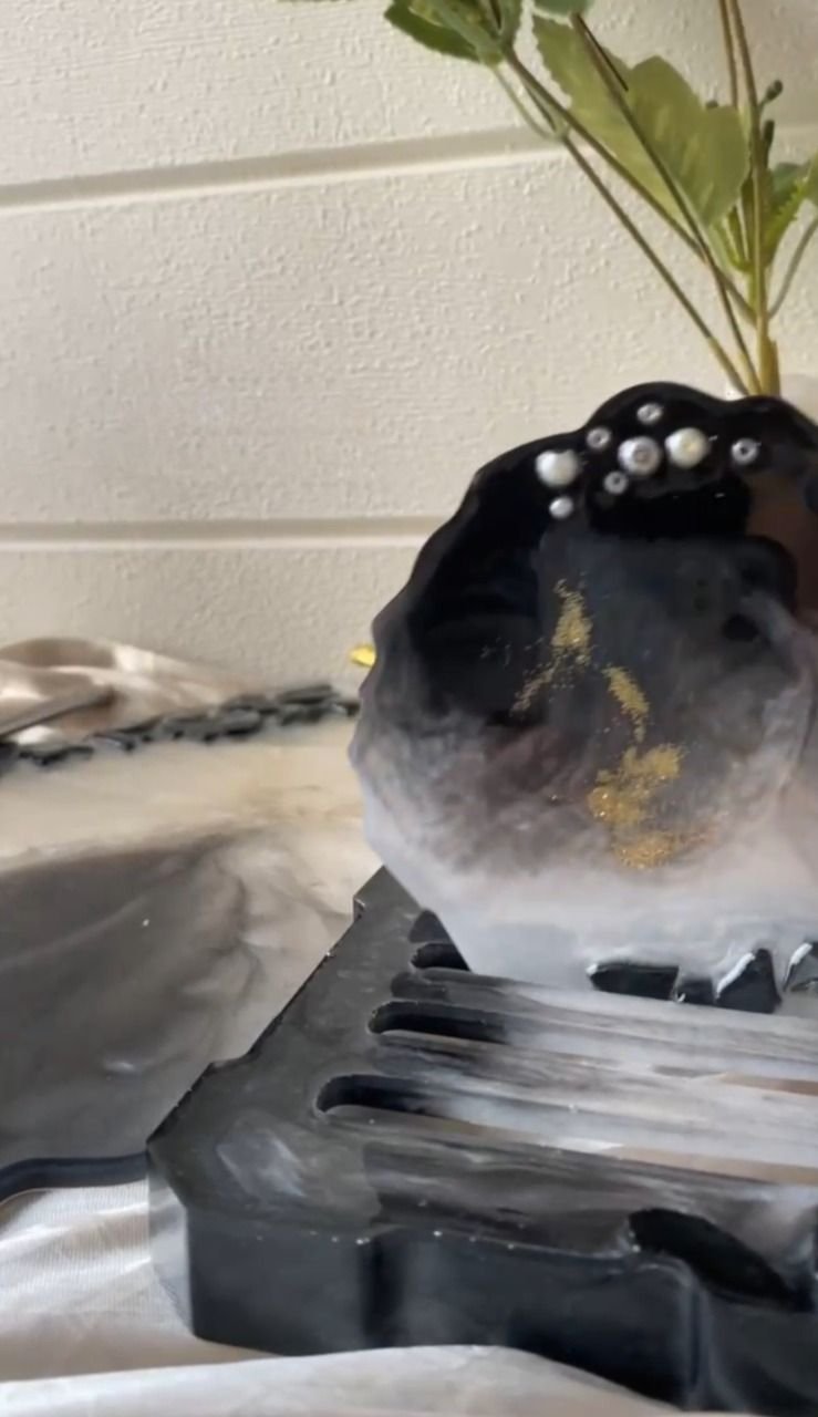 A round coaster of black and white marbled resin, veined with gold and set with a row of pearls along its top edge, standing upright in a matching slotted resin stand, with a plant behind it