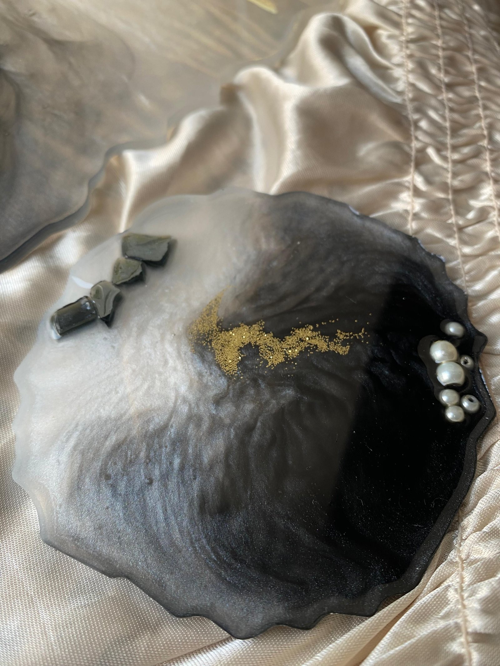 A round coaster of black and white marbled resin with an uneven rim, a vein of gold leaf running across it and a cluster of freshwater pearls set into one edge, lying on cream satin beside small dark stones