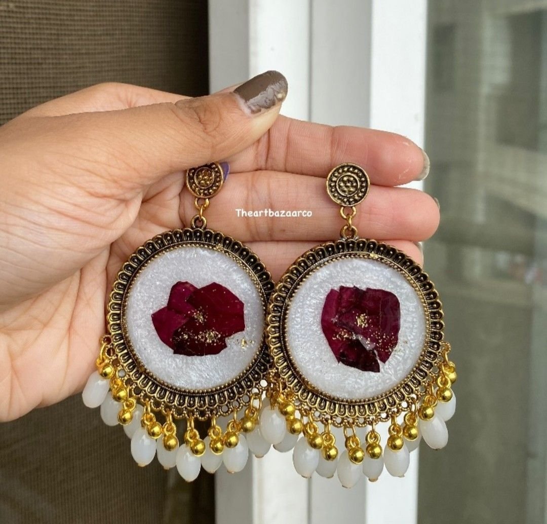 A pair of round jhumkas, each holding a deep maroon rose petal set into cream resin, framed in antique gold and fringed with white and gold beads, held in the hand