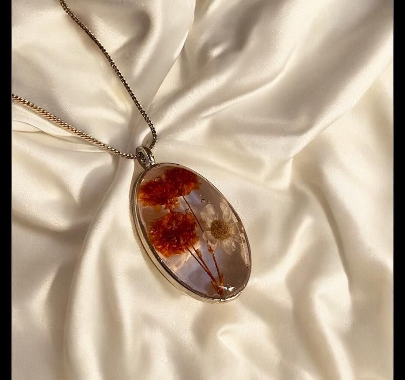 An oval pendant holding red and amber dried flowers in clear resin, on a fine chain over cream satin