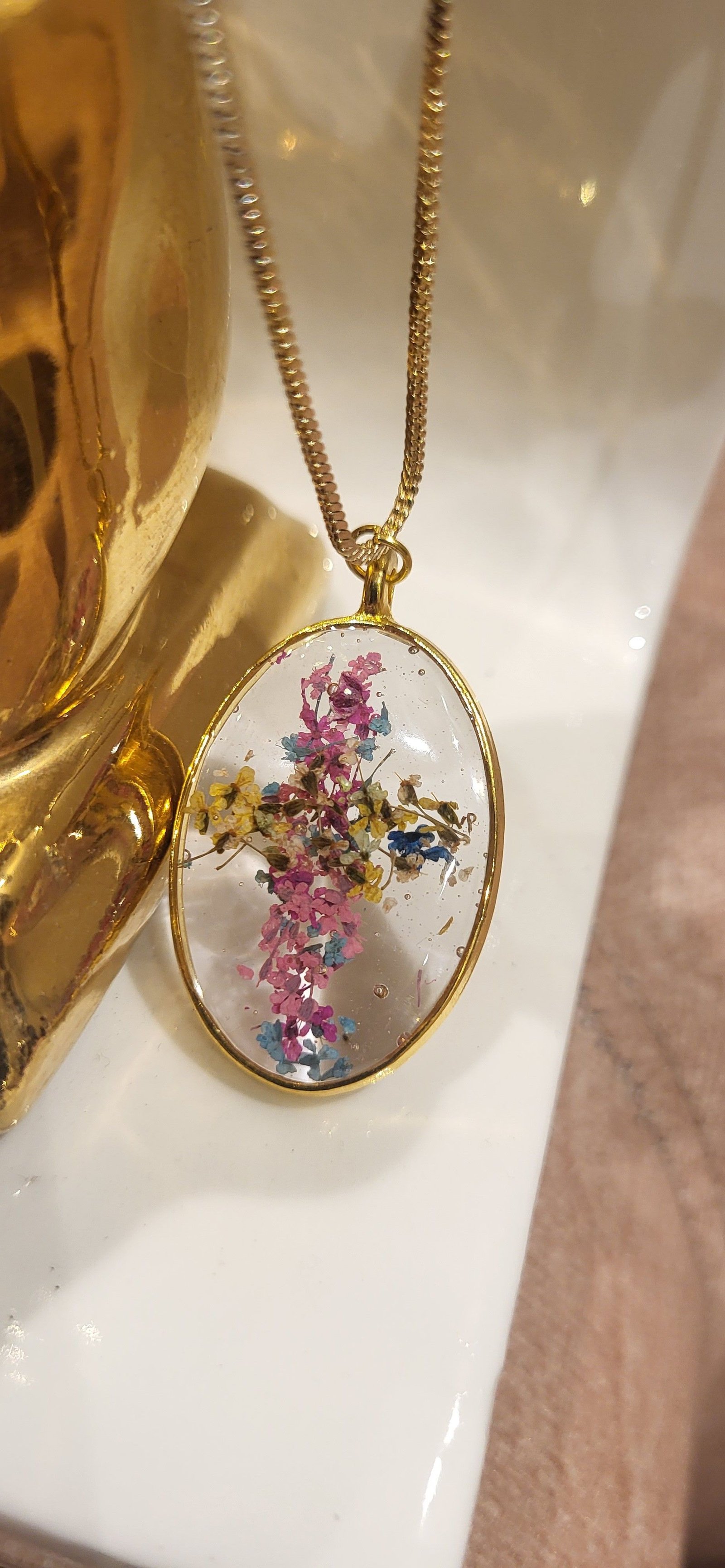An oval pendant in a slim gold frame, set with pressed pink, blue and yellow flowers suspended in clear resin, on a fine gold chain and resting against a gold vase