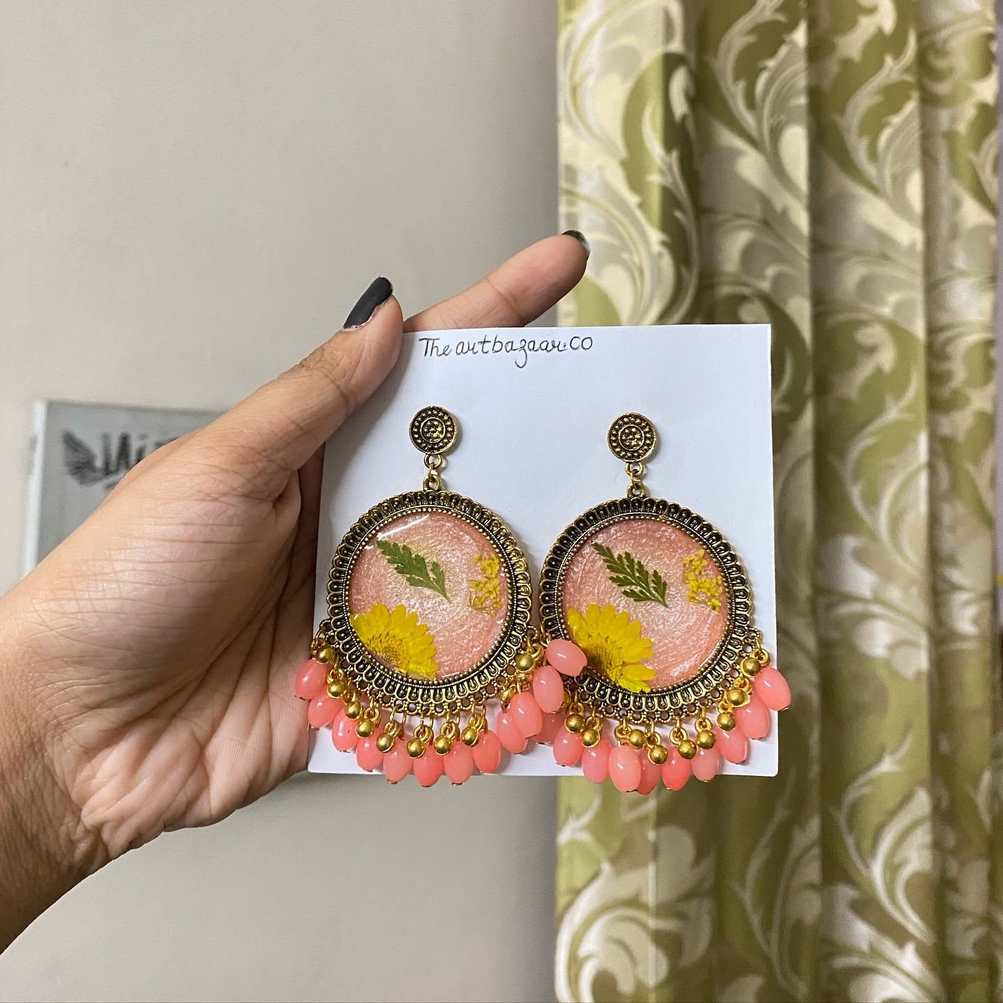 A pair of peach resin jhumkas, each set with a green fern frond and a small yellow daisy, framed in antique gold with a fringe of coral beads, pinned to a card handwritten with the maker’s name and held in the hand