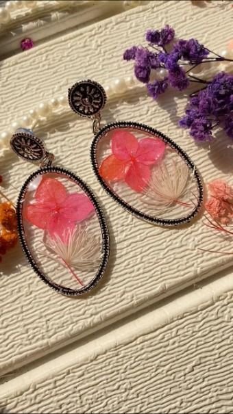 A pair of open oval earrings edged in small black beads, each holding a bright pink hydrangea petal and a pale spray of dried grass in clear resin, laid on textured cream with a strand of pearls and a sprig of purple statice behind them