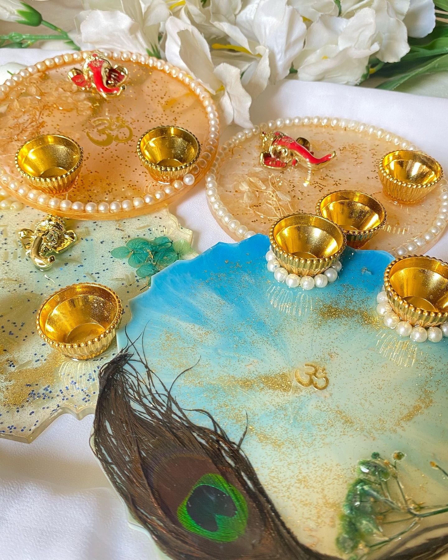 Three resin pooja thalis — two in peach and gold with pearl rims, Ganesha figures and brass diya cups, and one in sea blue and green with a peacock feather set beneath the surface