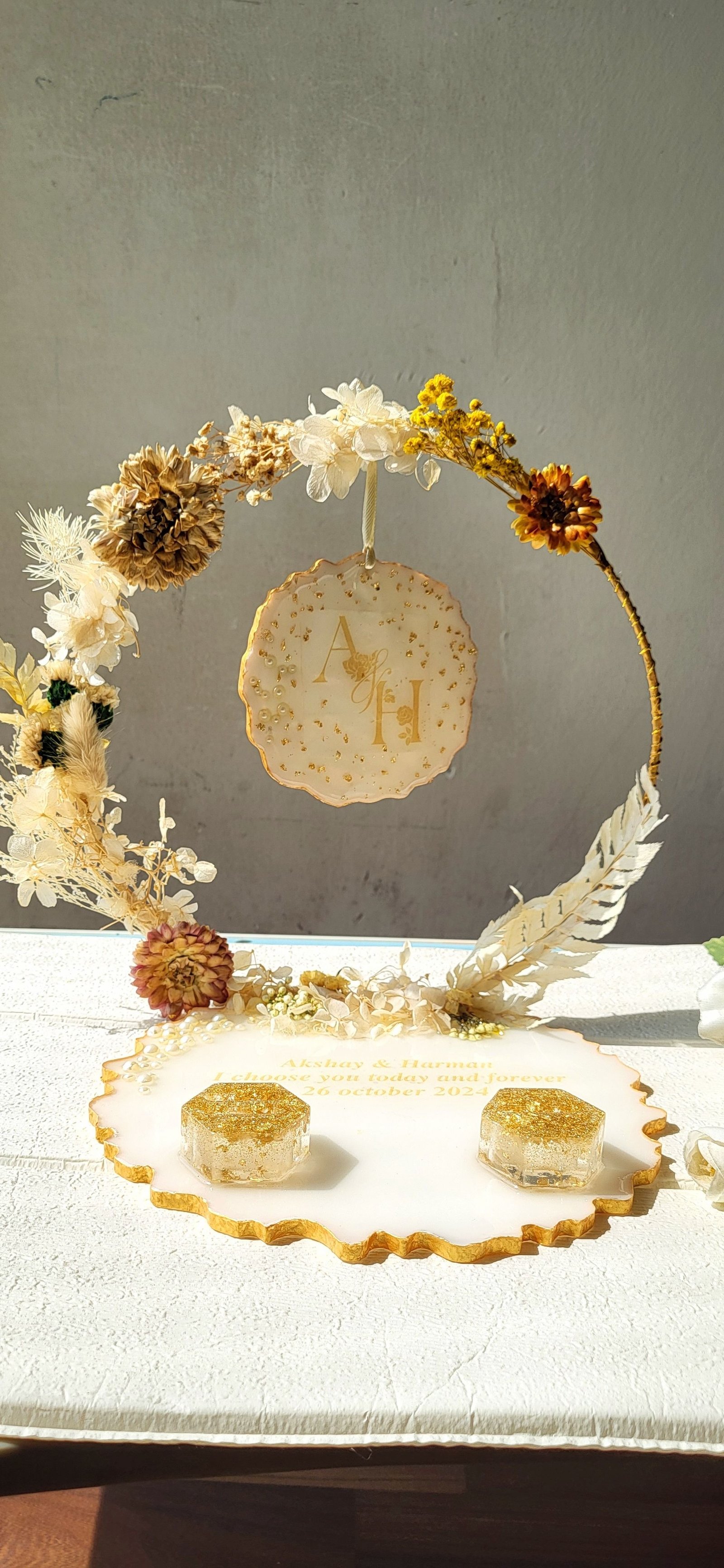 A gold hoop hung with preserved wedding flowers in cream, gold and rust, and a pale resin disc at its centre carrying the couple’s initials in gold leaf