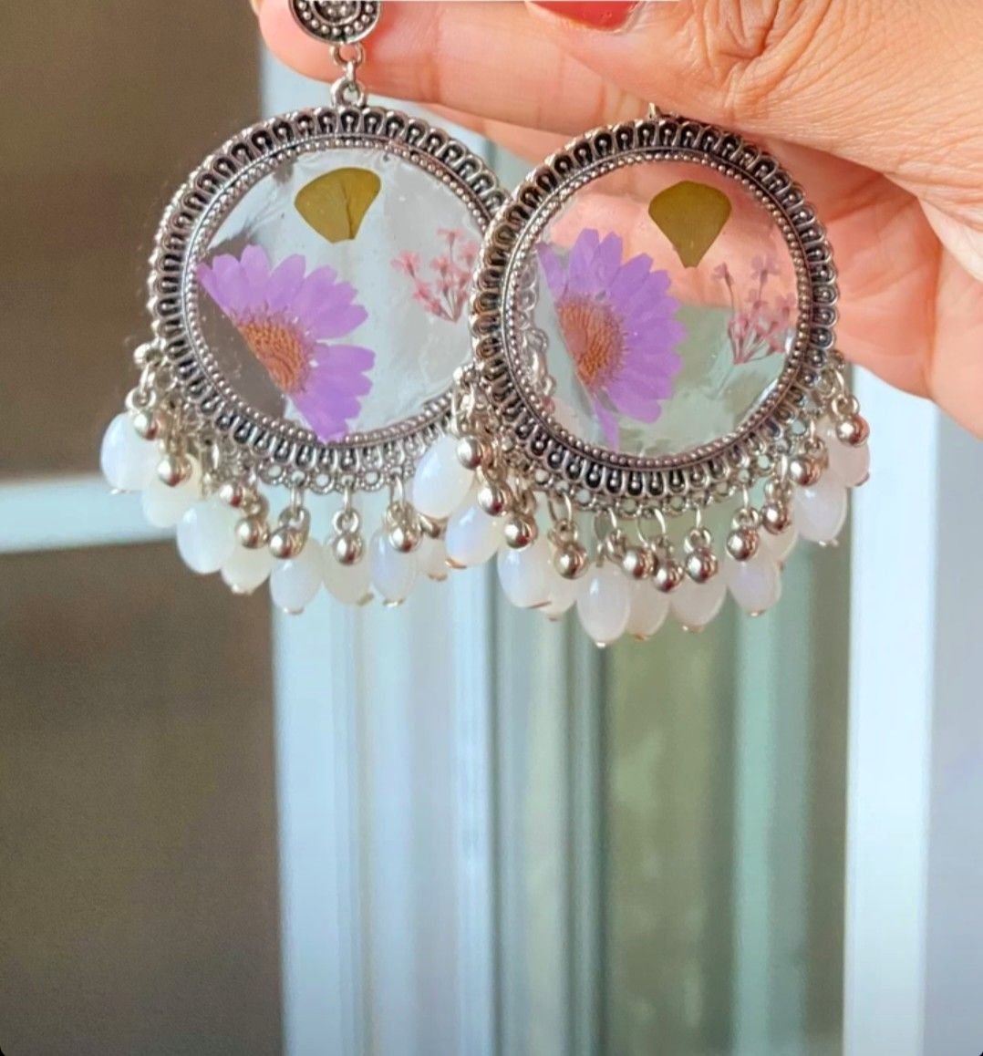 A pair of round jhumkas holding pressed lilac daisies under clear resin, framed in oxidised silver with a fringe of white beads, held up in the hand against a pale curtain