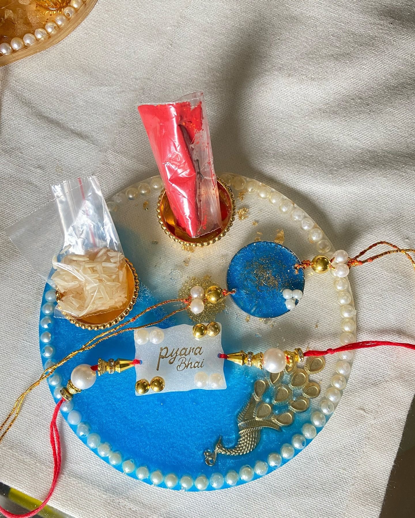 A round pooja thali poured in blue resin with a rim of pearls and a gold peacock, laid with two rakhis, a small card reading “pyara Bhai”, and gold katori bowls holding roli and rice