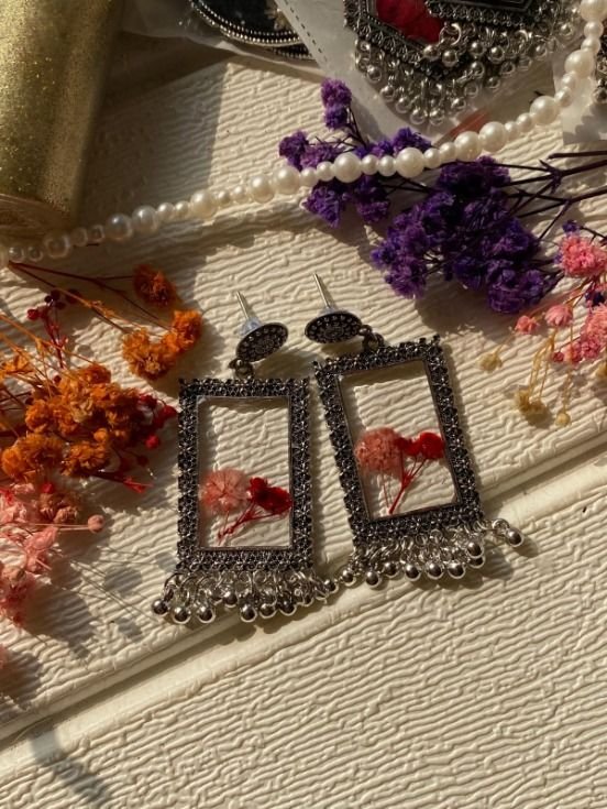 A pair of open rectangular jhumkas in oxidised silver with finely beaded borders, pink and red gypsophila set in clear resin inside each frame and a close silver ghungroo fringe below, laid on textured cream with a strand of pearls, a roll of gold ribbon and sprays of dried orange, pink and purple gypsophila around them