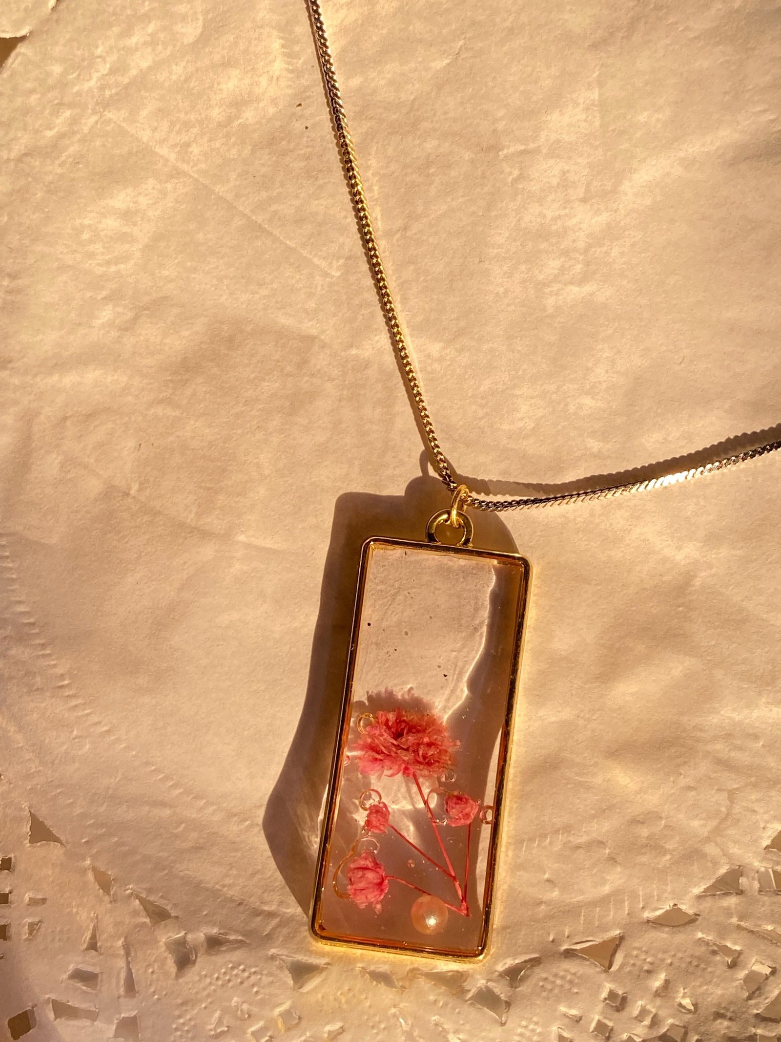A rectangular pendant of clear resin holding sprays of coral and pink blossom, framed in gold and hanging from a fine chain over cream lace