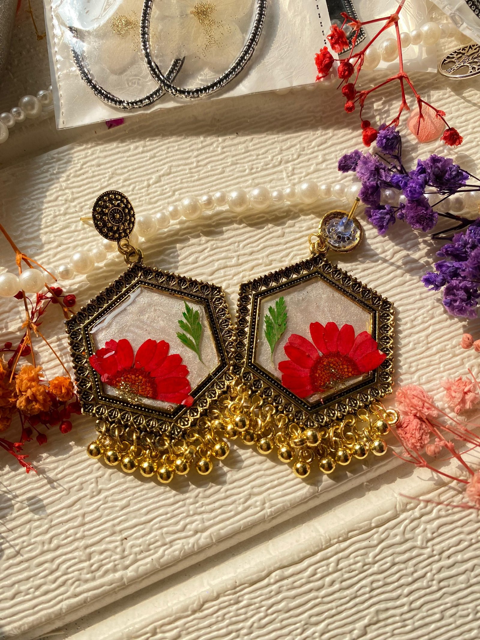 A pair of hexagonal jhumkas, each holding a whole red flower under clear resin, framed in antique gold with a heavy fringe of gold bells, lying on textured cream cloth with dried flowers around them