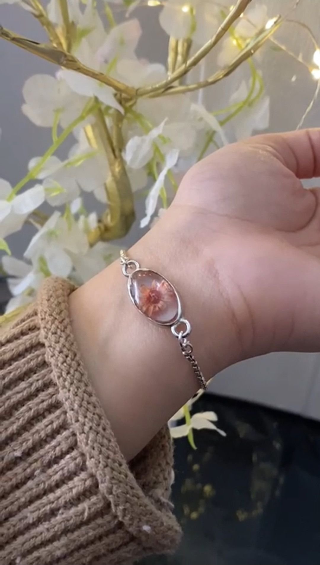 A fine silver chain bracelet with an oval resin centre holding a pink flower, worn on the wrist