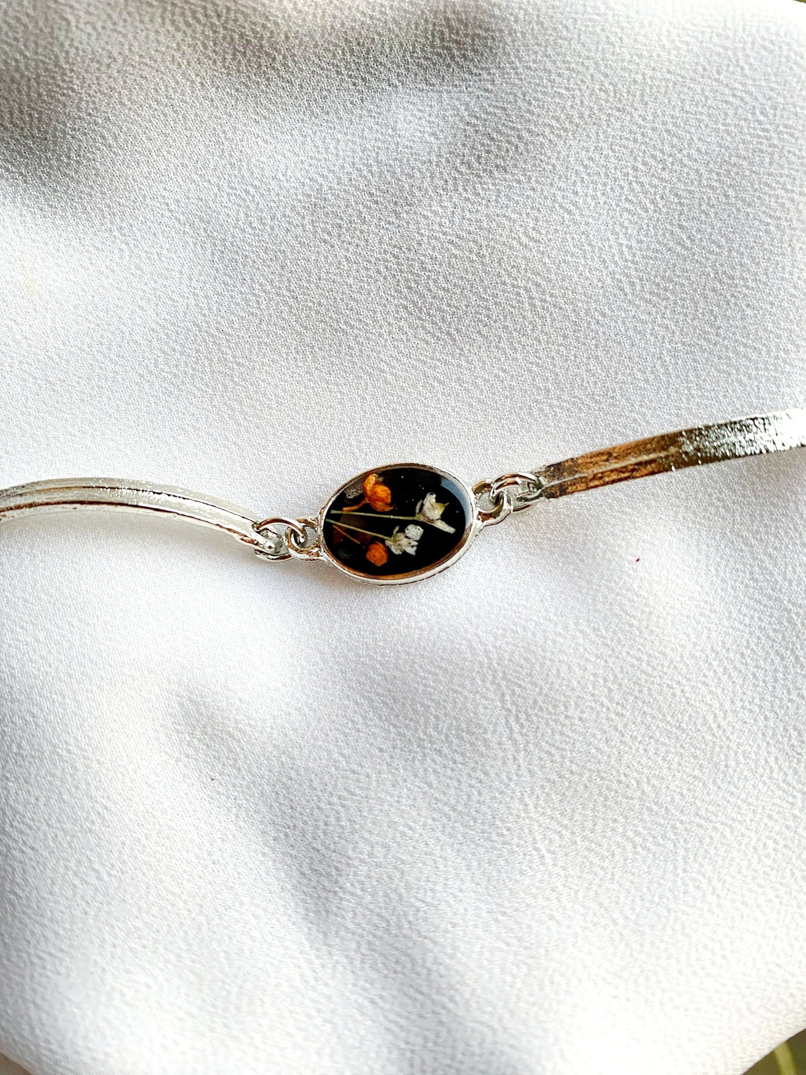 A gold bangle-style bracelet laid flat on white leather, its black resin oval holding orange and white flowers
