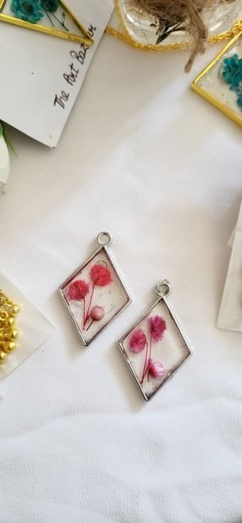 Two small silver-framed pendants lying on white cloth, one a rhombus and one a rectangle, each holding pink pressed blossoms on a fine stem under clear resin, with a card handwritten "The Art Bazaar" behind them