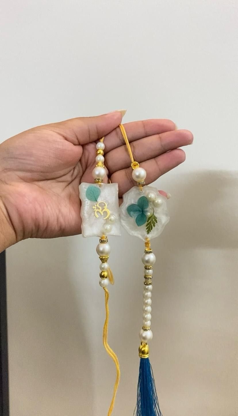 A pair of rakhis, each with a white resin square holding a blue flower, strung with pearls and finished with a blue silk tassel, held in the hand