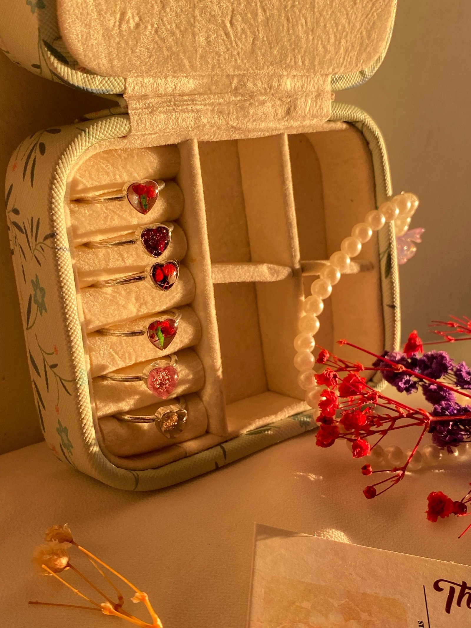 A row of resin rings set with small red and pink flowers, standing in a fabric-lined jewellery box beside dried blooms