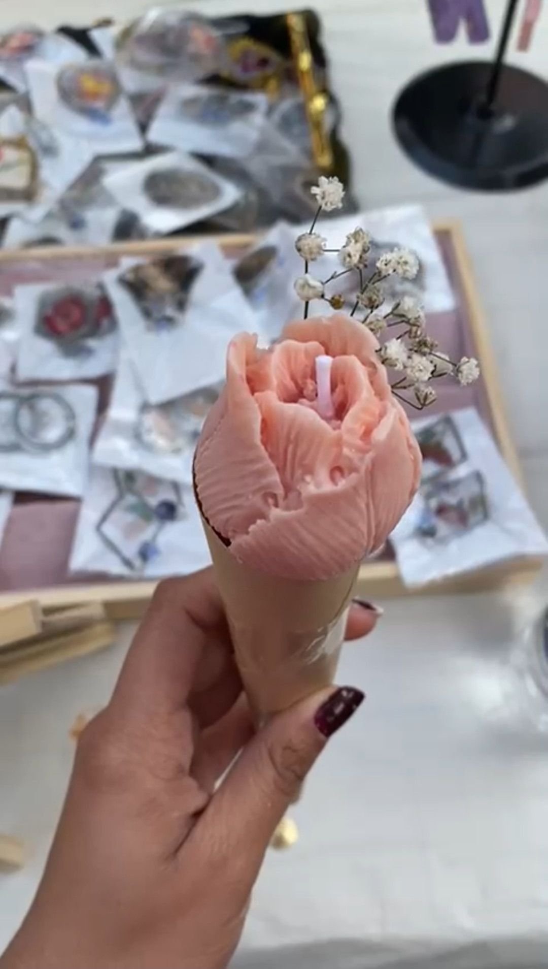 A pink wax rose wrapped in a paper cone, held in the hand at a market stall