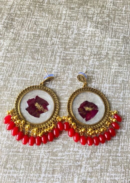 A pair of round antique-gold jhumkas laid on woven beige cloth, each holding deep magenta rose petals and scattered gold flakes in pale cream resin, with a fringe of red beads and small gold ghungroo below