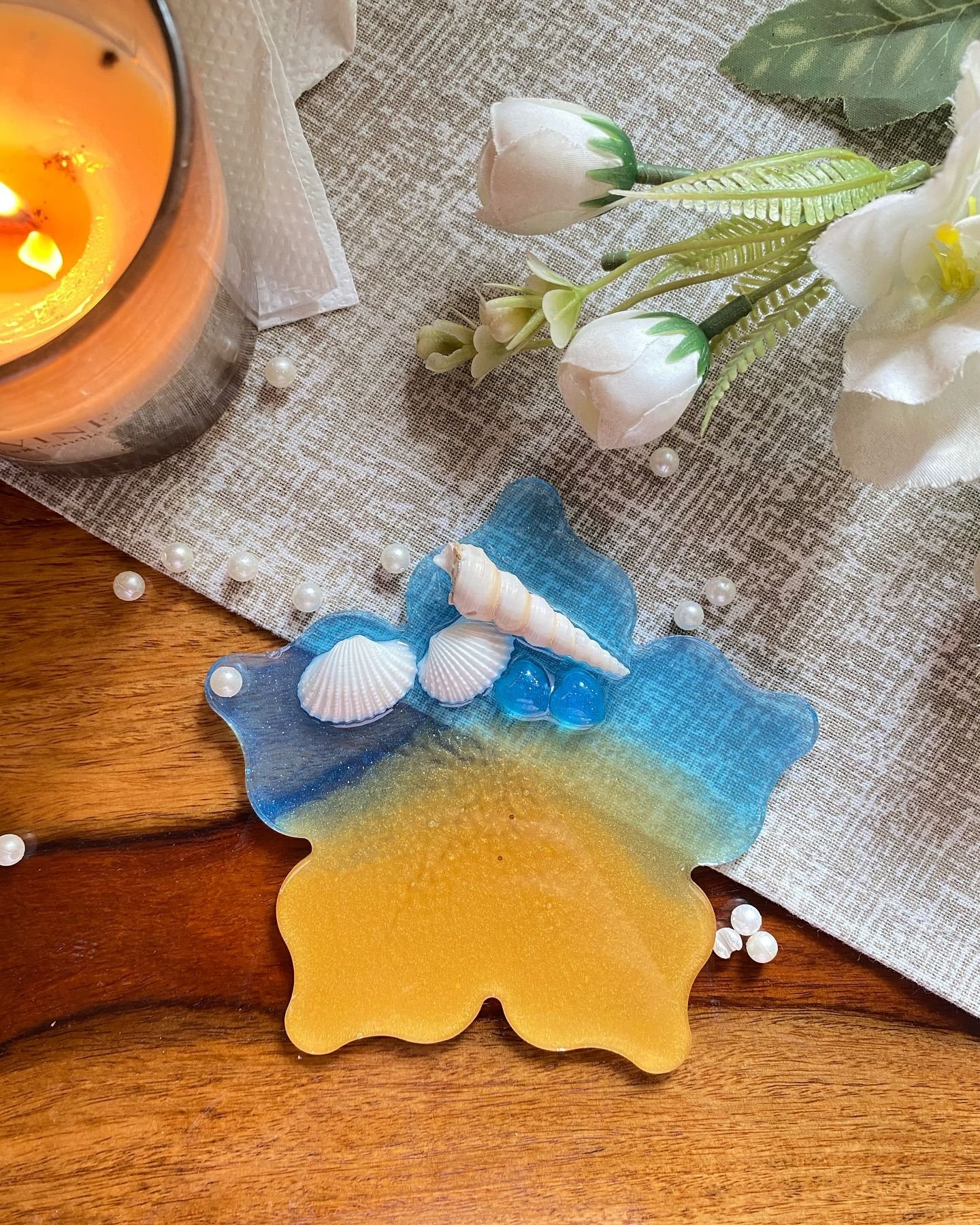 A resin coaster poured as a small beach scene — deep blue water shading into golden sand, with real seashells and blue glass beads set into the surface — resting on a wooden table beside a lit candle and white flowers