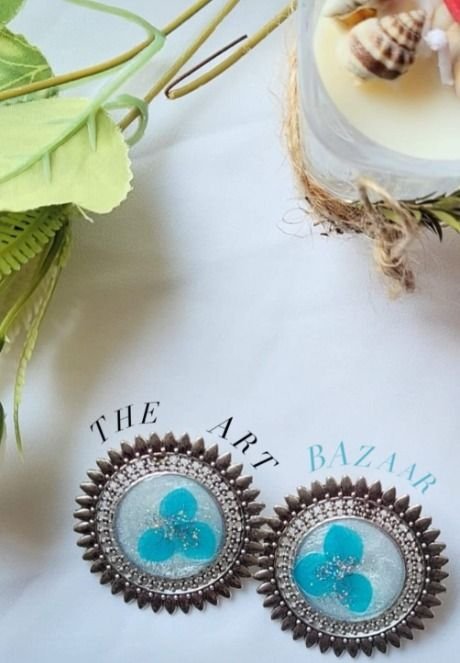 A pair of round studs in a spiked silver setting, pale resin holding three turquoise blossoms, on white cloth beside a candle and a shell