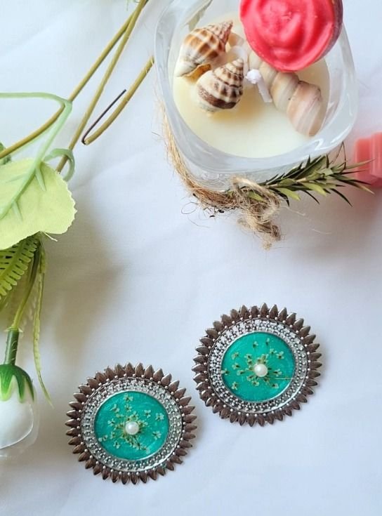 Round studs in the same spiked setting, this pair in sea-green resin with a single pearl and a spray of white gypsophila, photographed in daylight beside a candle and greenery