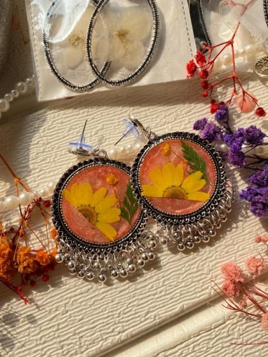 A pair of round oxidised silver jhumkas in low sun, each holding a yellow daisy and a green fern frond on a coral ground with a silver ghungroo fringe below, laid on textured cream among dried orange and purple flowers and a strand of pearls, another pair still in its sleeve behind them