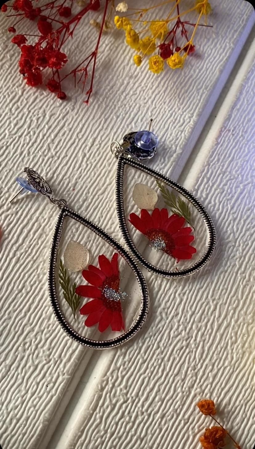 A pair of open teardrop earrings in blackened silver, each holding a single red daisy suspended in clear resin, lying on textured cream cloth