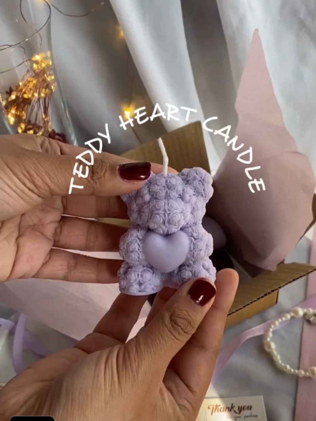 A lilac teddy bear candle held in the hand, wrapped in tissue with fairy lights behind