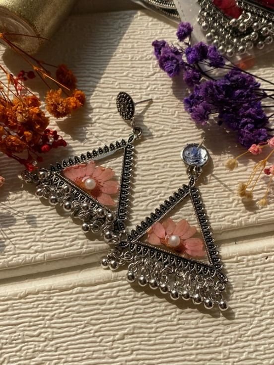 A pair of triangular jhumkas in oxidised silver, point down, each holding a pale pink pressed flower with a pearl at its centre under clear resin, silver ghungroo hanging from both lower edges, photographed in low sun on textured cream with dried orange and purple gypsophila around them