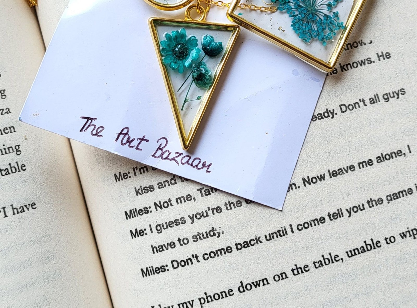 A gold-framed triangular pendant of teal pressed daisies resting on a small card handwritten "The Art Bazaar", laid across the pages of an open book, with a second gold pendant of teal flowers above it