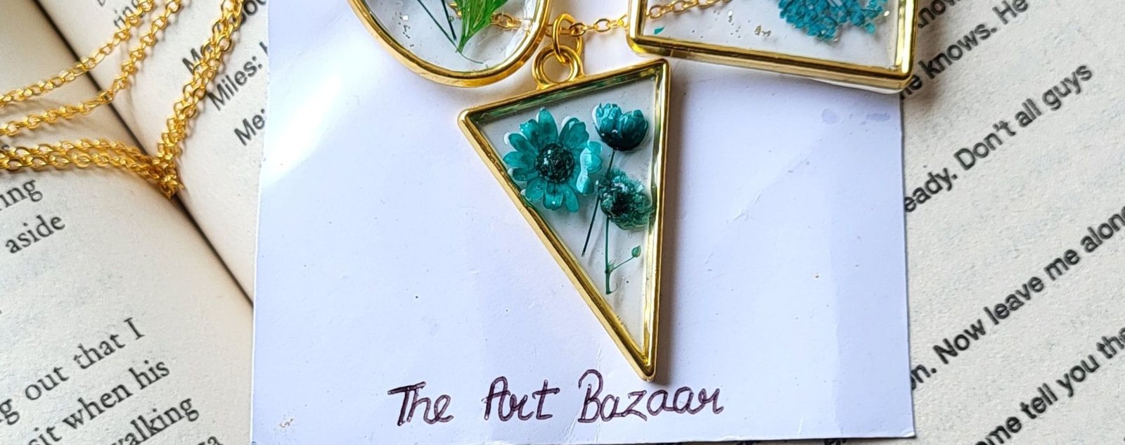 The same pendant and card photographed wider, a gold chain and an oval pendant holding a green fern lying along the top of the open book